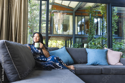Happy asian woman on couch with mug of coffee thinking at home photo