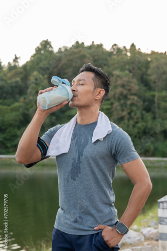 A happy, strong Asian man in sportswear is drinking a protein shake after a long run outdoors.