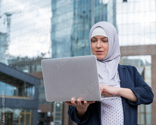 Thoughtful business woman in hijab and suit is holding a laptop outdoors.