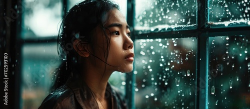 Asian woman alone, observing rain through home window.