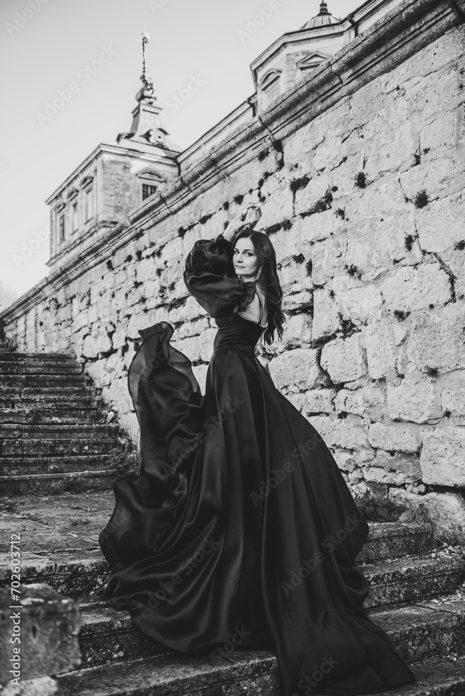 Woman in long dress walk on stairs near ancient palace at sunset. Model girl goes in street. Luxury bride near old Pidhirtsi Castle, Lviv, Ukraine. Stylish female. Back view. Black and white photo