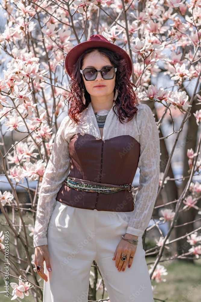 Magnolia park woman. Stylish woman in a hat stands near the magnolia bush in the park. Dressed in white corset pants and posing for the camera. Magnolia park woman. Stylish woman in a hat stands near the magnolia bush in the park. Dressed in white corset pants and posing for the camera.