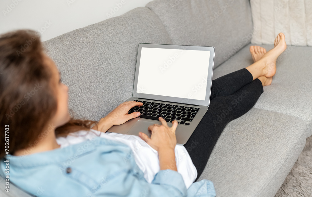 Fototapeta premium Over the shoulder view laptop with blank screen on laps of woman. Woman uses her laptop while relaxing at home on sofa.