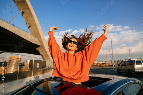 Canvastavla An exuberant emotional young woman with excitement as she leans out of the car sunroof with outstretched arms on the city street