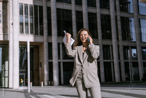 Happy businesswoman laughing on the phone, clutching documents, in front of a modern building