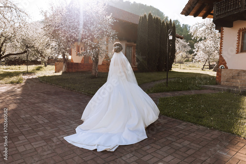 The bride has her shoulders turned to the camera, walking along a stone path. A long train. Magnificent dress with long sleeves, open bust. Spring wedding