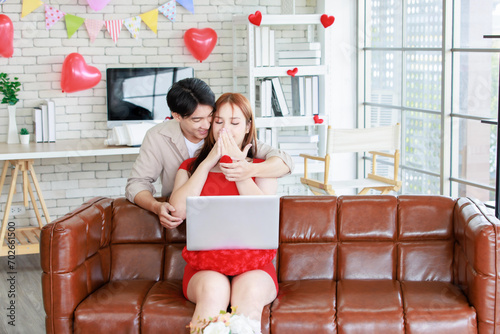 Asian young handsome male boyfriend standing smiling behind giving red wrapped present gift box beautiful female girlfriend sitting on sofa using laptop computer living room celebrating valentine day