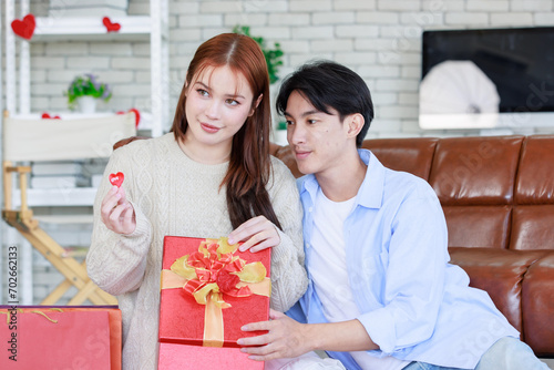 Asian young beautiful female girlfriend sitting smiling opening unboxing red present gift wrapped box on cozy sofa from handsome male boyfriend in romantic decorated living room on valentine day