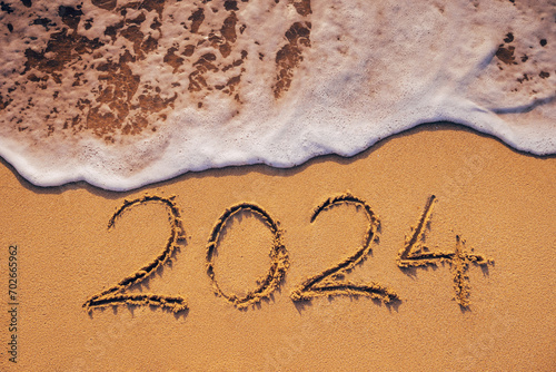 Happy New Year 2024 text written on the beach sand and ocean wave washing the sea shore