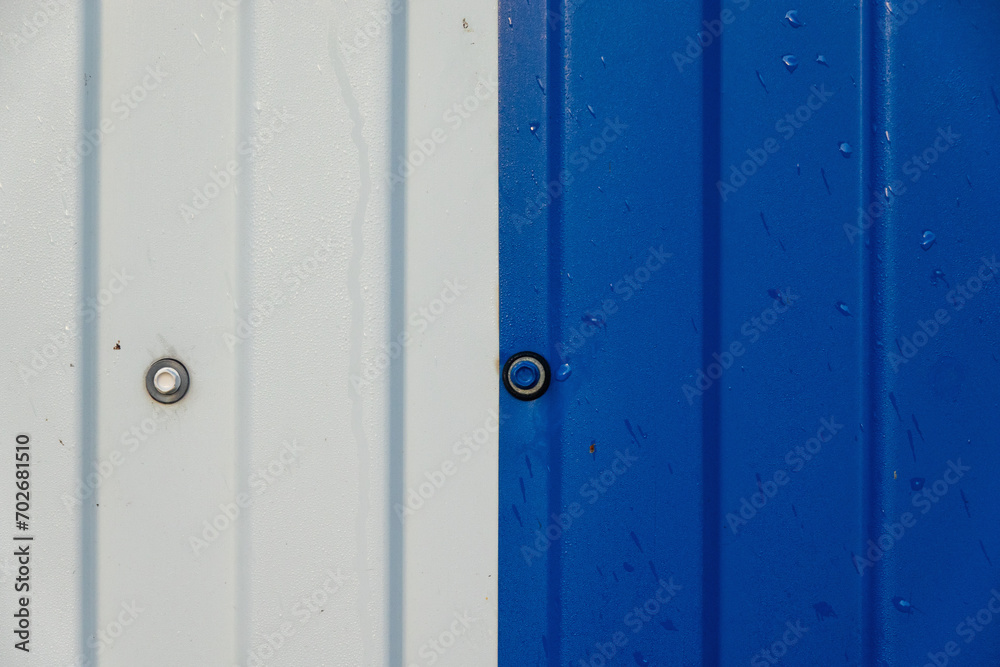 Close-up of a fragment of a metal fence. In the middle of the frame there is a connection of two steel sheets - white and blue. There are mounting screw heads and raindrops. Background. Close-up of a fragment of a metal fence. In the middle of the frame there is a connection of two steel sheets - white and blue. There are mounting screw heads and raindrops. Background.