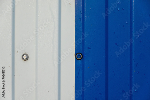 Close-up of a fragment of a metal fence. In the middle of the frame there is a connection of two steel sheets - white and blue. There are mounting screw heads and raindrops. Background.
