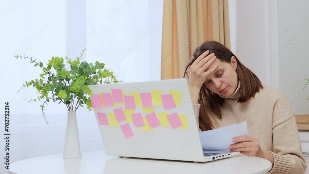 Sad worried Caucasian woman using laptop holding papers paying bills sitting at home in kitchen going over her utility bills thinking about financial problems Sad worried Caucasian woman using laptop holding papers paying bills sitting at home in kitchen going over her utility bills thinking about financial problems