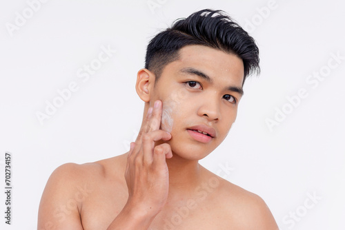 A young asian man applies exfoliating facial scrub cream to his face using his fingers. Skin care and hygiene concepts. Isolated on a white backdrop.