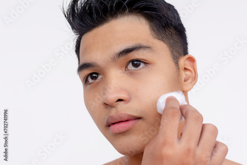 A young asian man using a cotton ball dipped in astringent or skin toner to cleanse his face. Using a dabbing motion. Skin care and hygiene concepts. Isolated on a white backdrop. photo