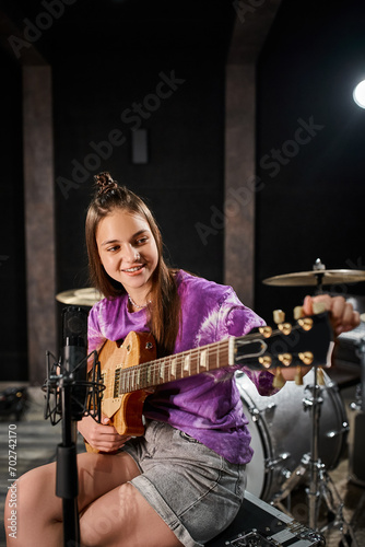 cheerful teenage guitarist in casual vivid attire playing guitar in front of microphone in studio