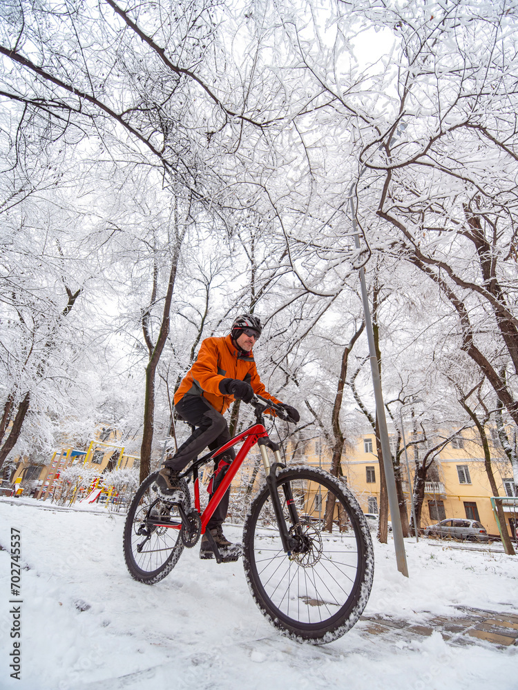 A guy in an orange jacket rides a bicycle through a winter city among snow covered trees. Active lifestyle in winter