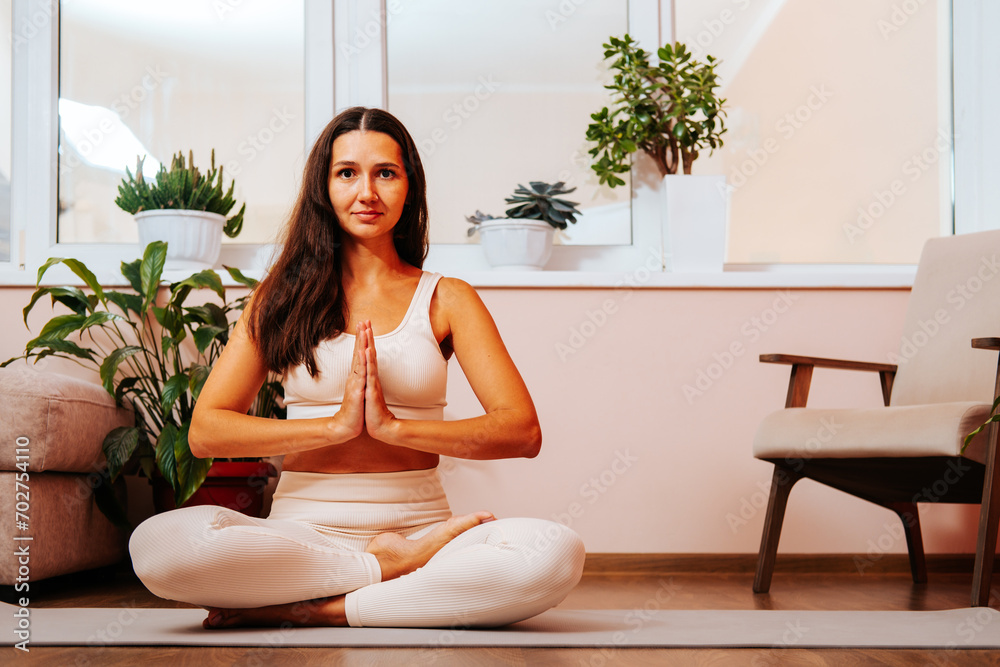 Beautiful young brunette woman is sitting in lotus position with her hands in namaste on yoga mat at home in living room. Beautiful young brunette woman is sitting in lotus position with her hands in namaste on yoga mat at home in living room.