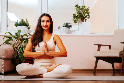 Beautiful young brunette woman is sitting in lotus position with her hands in namaste on yoga mat at home in living room.
