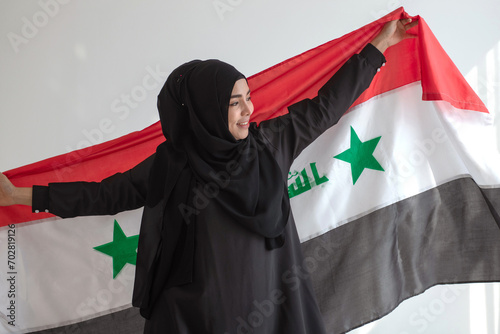 Happy Muslim woman wears black Hijab Muslim traditional clothes holding Irag flag, sunlight and shade on white wall, Muslim woman rights concept photo