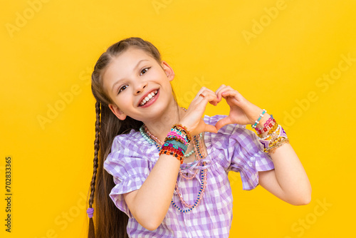 The little girl shows her heart with her fingers and smiles happily. The child put on a lot of beaded bracelets. Beading for children. Yellow isolated background.