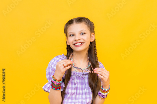 The child put on a lot of bracelets made of beads and beads. A happy little girl is enjoying a beaded ornament. Beading for children. Isolated yellow background.