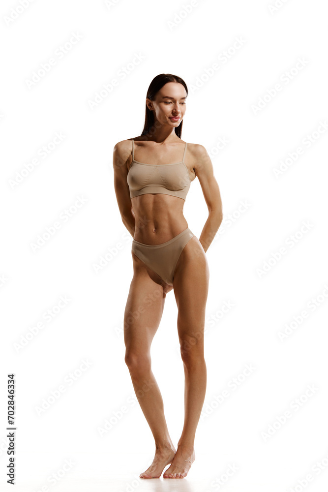 Brunette young woman with slim muscular body standing in beige cotton underwear against white studio background. Concept of female beauty, body and skin care, cosmetology, sport, health