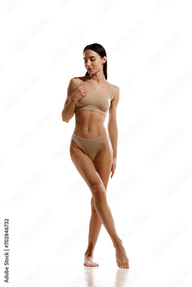 Fototapeta premium Full-length image of slim beautiful brunette woman with relief body standing in beige cotton underwear over white studio background. Concept of beauty, body and skin care, cosmetology, sport, health