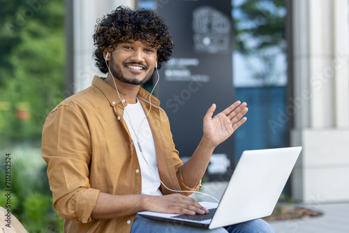 Portrait of a young Muslim man wearing headphones, holding a laptop on his lap, sitting on a bench in a city street, smiling and waving at the camera