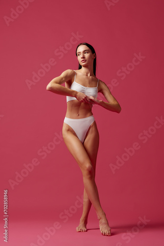 Body-positivity. Full-length image of young beautiful woman with fit, slim, healthy body in white cotton underwear against pink studio background. Concept of beauty, body and skin care, cosmetology