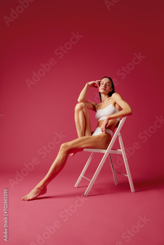 Full-length image of young woman with slim fit healthy body sitting on chair in white cotton underwear against pink studio background. Concept of female beauty, body and skin care, spa, sport, health