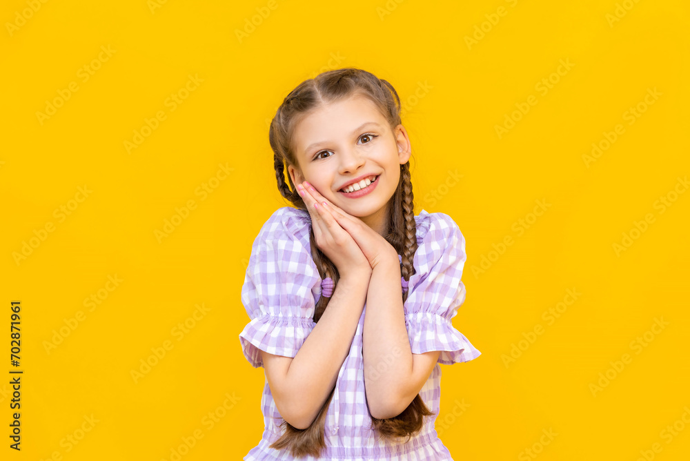 A charming little girl, adoringly clasping her hands in front of her face. The child is smiling blissfully on a yellow isolated background. A happy young girl. A charming little girl, adoringly clasping her hands in front of her face. The child is smiling blissfully on a yellow isolated background. A happy young girl.
