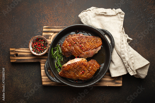 Two roasted duck breast fillets with crispy skin, with pepper and rosemary, top view in black cast iron pan with knife, dark brown concrete rustic background.