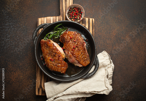 Two roasted duck breast fillets with crispy skin, with pepper and rosemary, top view in black cast iron pan with knife, dark brown concrete rustic background.