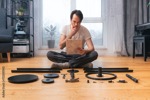 A man is intently studying the instructions for self-assembly of furniture, sitting cross-legged on the floor and leaning his head on his hand, among neatly laid out parts and tools.