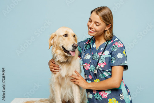 Young smiling happy veterinarian woman she wear uniform stethoscope heal exam hug cuddle embrace retriever dog isolated on plain pastel light blue background studio portrait. Pet health care concept.