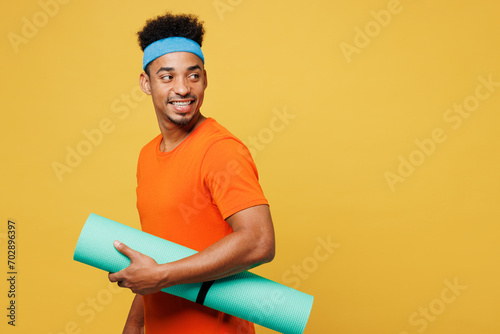 Sideways young fitness trainer sporty man sportsman wear orange t-shirt hold in hand yoga mat look aside area spend time in home gym isolated on plain yellow background. Workout sport fit abs concept.