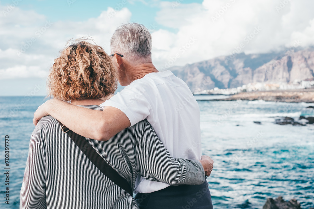 Fototapeta premium Rear view of carefree bonding senior couple embrace on the seashore looking at horizon over water, two elderly caucasian people stay together expressing love and tenderness