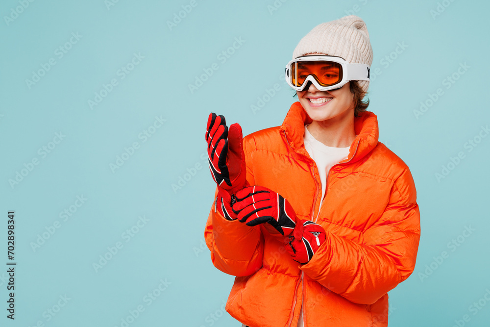 Young smiling happy skier woman she wear warm padded windbreaker jacket hat ski goggles mask put on red gloves travel rest spend weekend winter season in mountains isolated on plain blue background. Young smiling happy skier woman she wear warm padded windbreaker jacket hat ski goggles mask put on red gloves travel rest spend weekend winter season in mountains isolated on plain blue background.