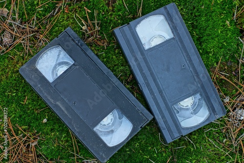 two old black dirty plastic video cassettes with magnetic tape lie on green moss on the street during the day