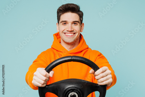 Young smiling happy man he wears orange hoody casual clothes hold steering wheel driving car look camera isolated on plain pastel light blue cyan color background studio portrait. Lifestyle concept.