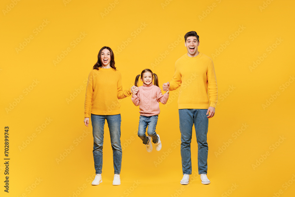 Full body smiling happy fun young happy parents mom dad with child kid girl 7-8 years old wear pink sweater casual clothes hold hands jump high isolated on plain yellow background. Family day concept.