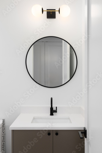 A bathroom detail with a brown cabinet, white marble countertop, and a black and gold light fixture above a black circular mirror.