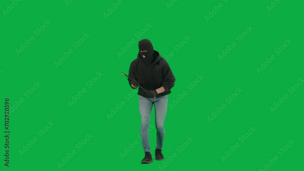 Portrait of thief on chroma key green screen background. Man robber in balaclava and hoodie walking holding crowbar in hands looks around. Portrait of thief on chroma key green screen background. Man robber in balaclava and hoodie walking holding crowbar in hands looks around.