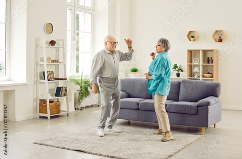 Full length photo of happy senior elderly family couple wife and husband dancing in the living room at home. Smiling retired man and woman having fun, enjoying spending time together on retirement. photo