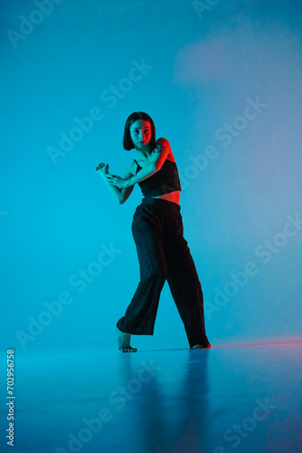 Young woman in black pants and top poses in studio against neon blue background. The dancer demonstrates the choreography elements of an experimental hip hop style dance. photo