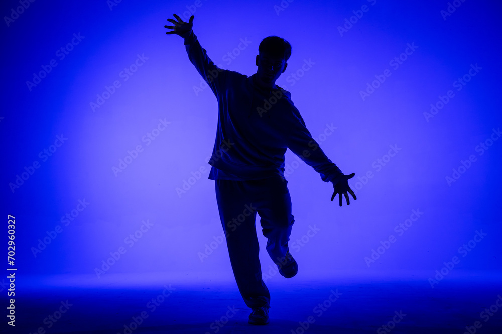 Silhouette of guy in casual clothes dancing elements of hip hop in studio with blue light. Dancer demonstrates body plasticity. Full height. Modern street choreography.