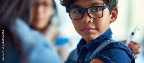A young Latino boy, 6, with glasses and a blue shirt, displays his bandaged arm after receiving a Covid-19 vaccine in the post-Coronavirus era. photo