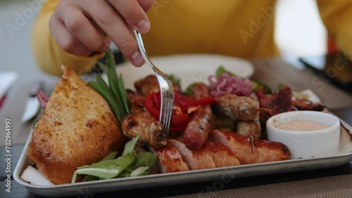 Unrecognizable traveler touches large Balkan dish with assorted meats and vegetables with fork in local authentic bar. Closeup of grilled meat, cevapcici and Serbian pljeskavica in flatbread. photo