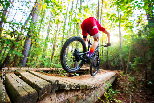 Mountain biker in a red jersey bounding over a wooden bridge on a trail in sunny woods, wide angle shallow focus photo
