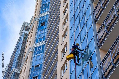 Bottom view of industrial mountaineer man laborer in work uniform washing glazing, hanging from building. Male rope access worker during high-rise job. Industry urban works concept. Copy ad text space photo